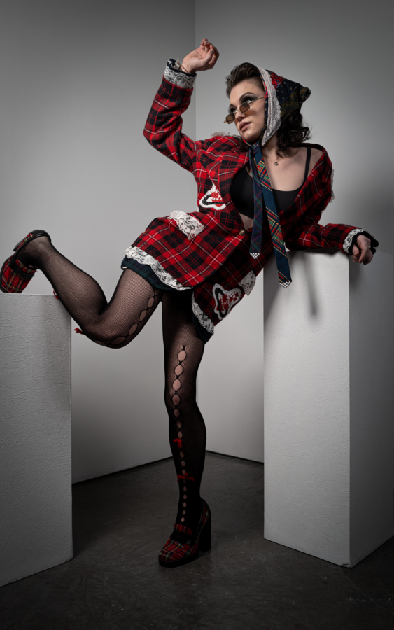 The NYFW model is posed in a blazer dress that is red and black plaid. They are wearing fishnet stockings and closed toe heels.